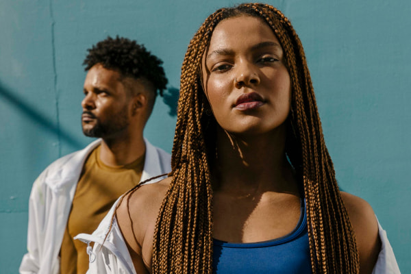 Two people, one person with dreaded hair in the front and looking to the camera, the person behind is looking to the right.